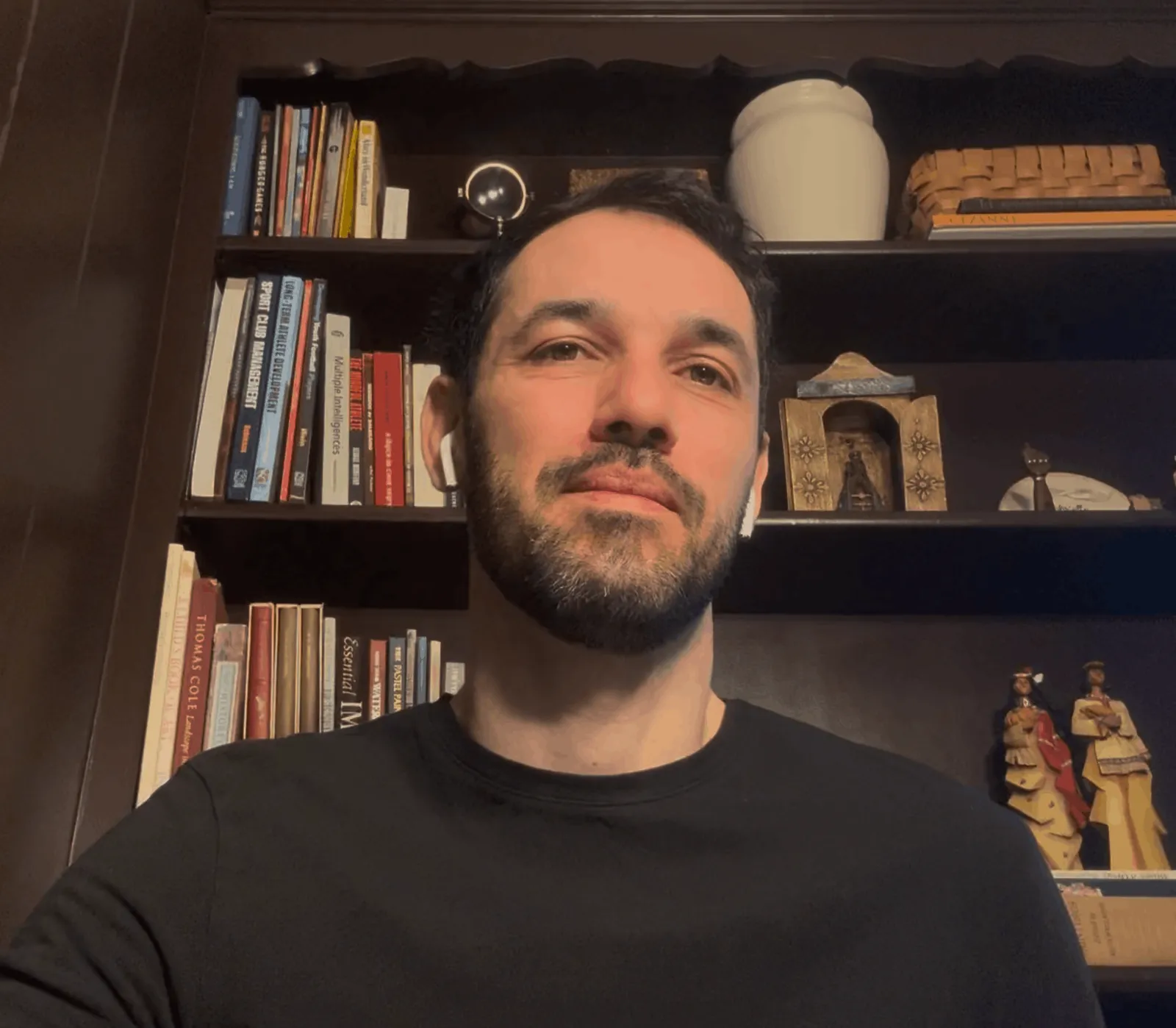 Man with a beard wearing white earbuds and a black shirt, sitting in front of a dark wooden bookshelf filled with books and decorative items.